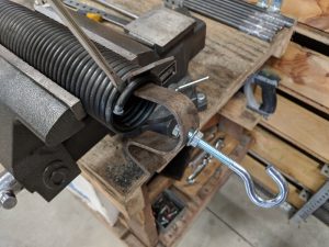 A garage door spring that has a clip with a hole in it and an eye bolt attached.  