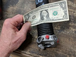 An image of a hand trying to place a one-dollar bill in between standard torsion spring coils. However, the bill does not fit in between the coils.